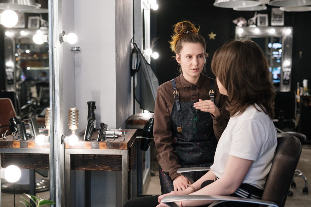 A lesbian woman is depicted in a friendly and respectful conversation with a queer hairstylist. She is enthusiastically describing the specific hairstyle she desires, using gestures to emphasize her points. The hairstylist is attentively listening and nodding, conveying understanding and readiness to create the requested style.