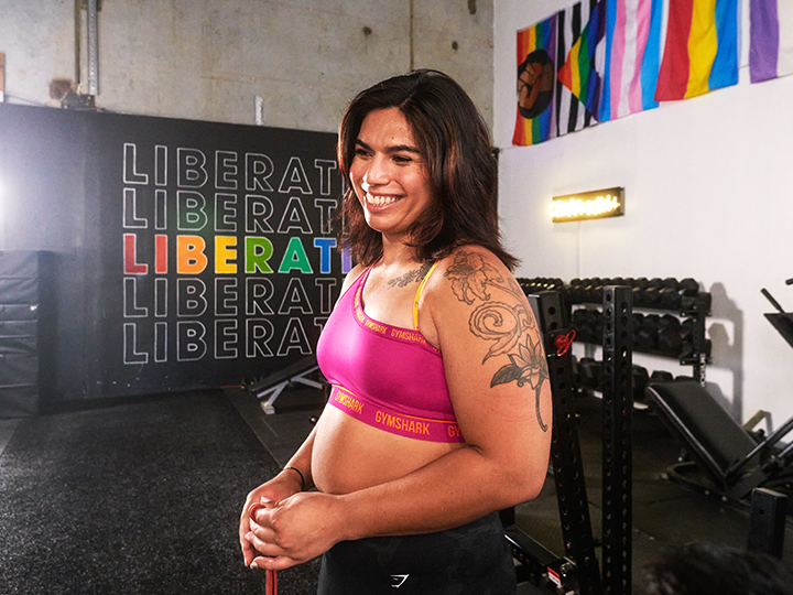 Transgender powerlifter and lesbian athlete Angel Flores stands in a pink sports bra in a gym with pride flags and a sign that reads “Liberation” behind her.