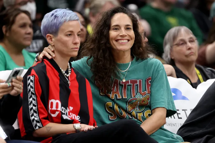Queer athlete couple Sue Bird and Megan Rapinoe pose together at a basketball game, they are both dressed in iconic sports jerseys.