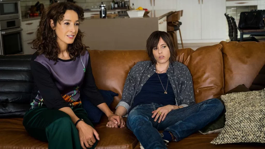 An image of Kate Moennig and Jennifer Beals playing Shane and Bette from the L Word Gen Q. They are holding hands on a brown leather couch. 
