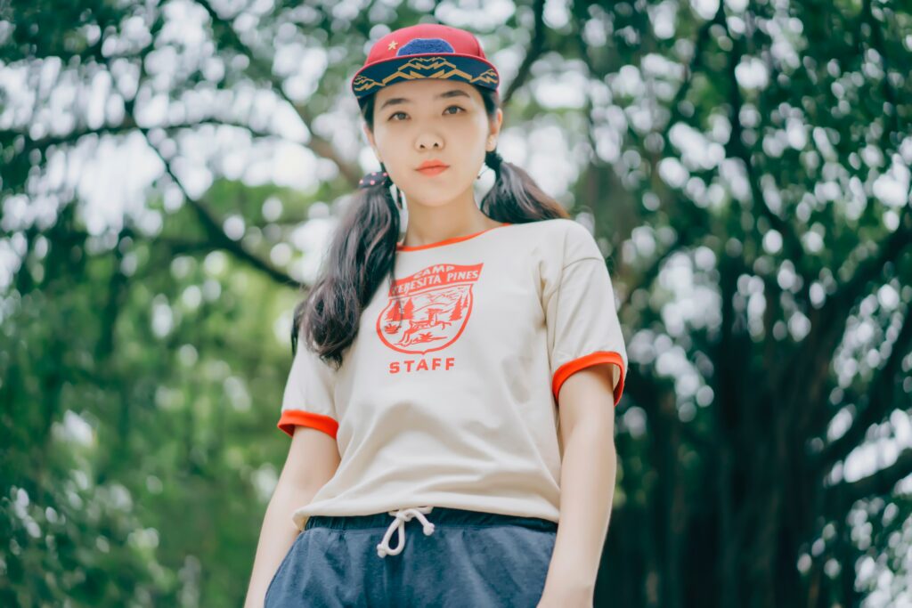 An Asian futch top with pigtails standing in a forest with a white and red ringer tee on and a snap back staring confidently into the camera.