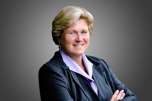 Gail Koziara Boudreaux posing with a smile and arms crossed on a gray background.