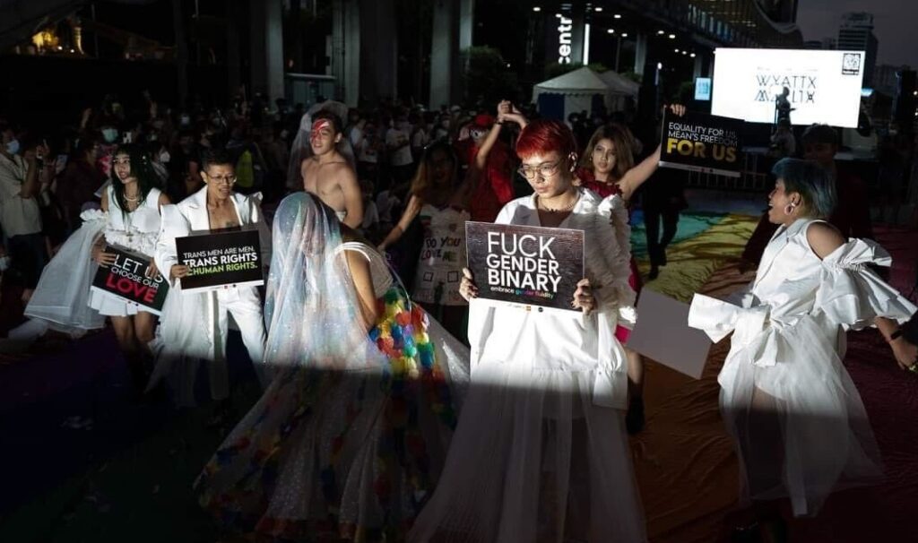 Image of a diverse group of LGBTQIA+ individuals proudly marching on a vibrant rainbow flag. They are holding posters filled with empowering messages, symbolizing unity and rights for the LGBTQIA+ community. Prominently displayed among these posters is the logo of HER, showcasing their alliance and support for the event.