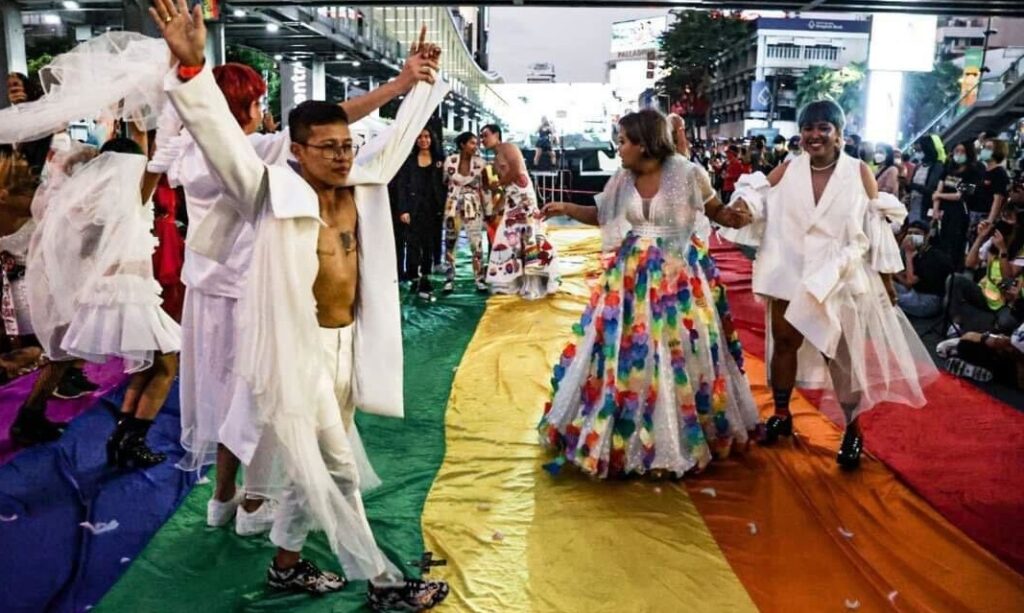A group of LGBTQIA+ individuals joyfully walking on a vibrant rainbow flag in the streets of Bangkok, symbolizing pride and diversity.