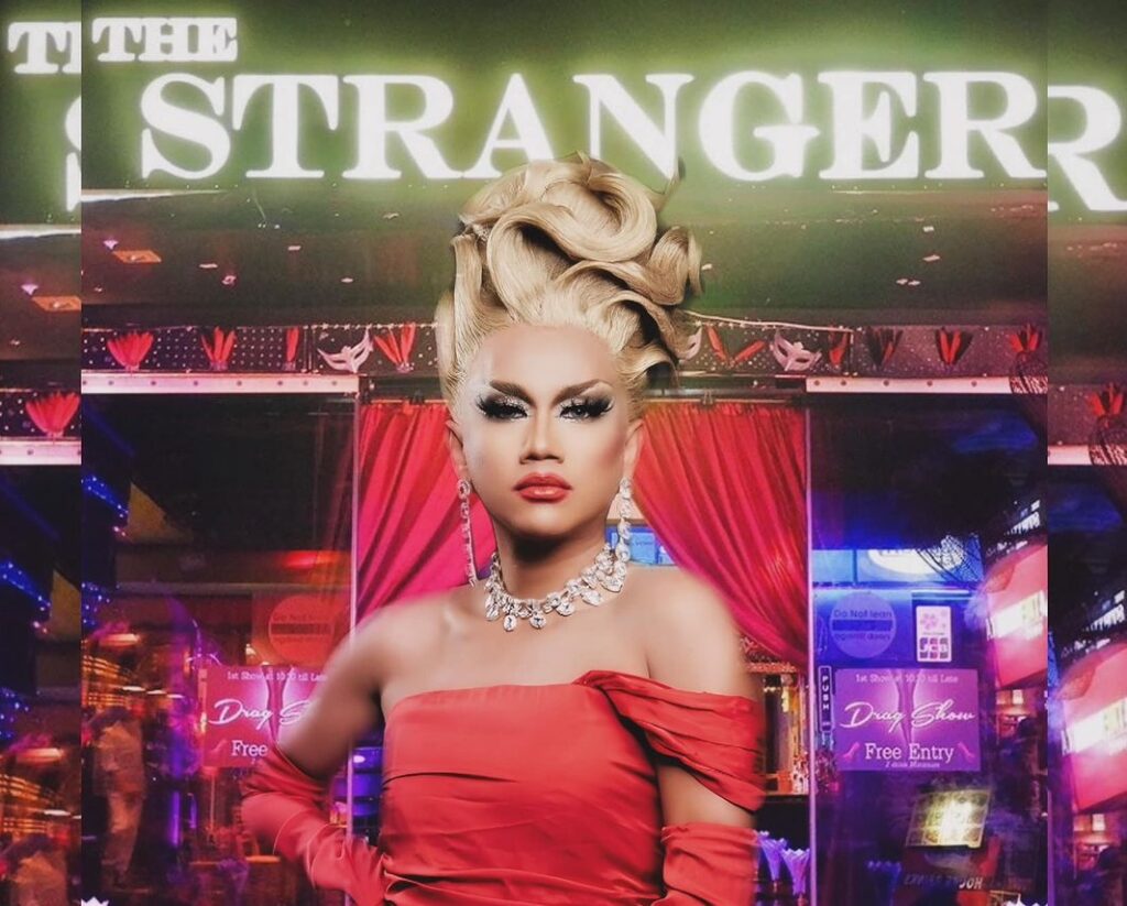 Elegant and commanding, M Stranger FOX dazzles in a red outfit, complemented by a glistening diamond necklace and a chic blonde wig, striking a confident pose.
