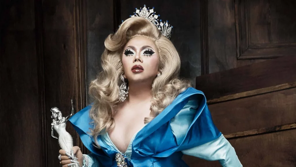 Assadayut Khunviseadpong, known as 'Natalia Pliacam,' elegantly dressed in a blue costume with a blonde wig, holding a metallic grey female sculpture, gazing thoughtfully out of frame, embodying both grace and artistic expression.
