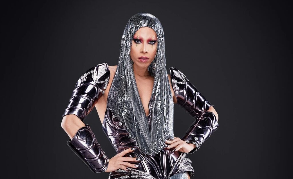 Pangina Heals, in her drag persona, exudes confidence and style in a shimmering grey outfit and wig, striking a bold pose with her hand on her hip and a fierce gaze into the camera.