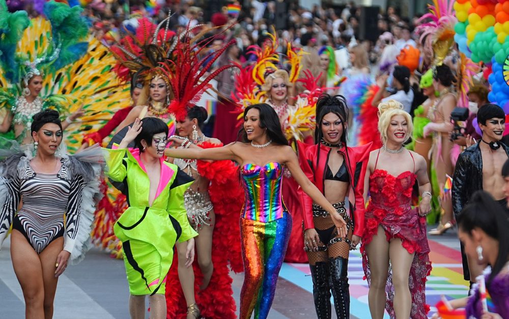 A lively scene featuring members of the LGBTQIA+ community and fabulous drag queens strutting their stuff at a Pride event in Bangkok, all dressed in vibrant rainbow-themed outfits.