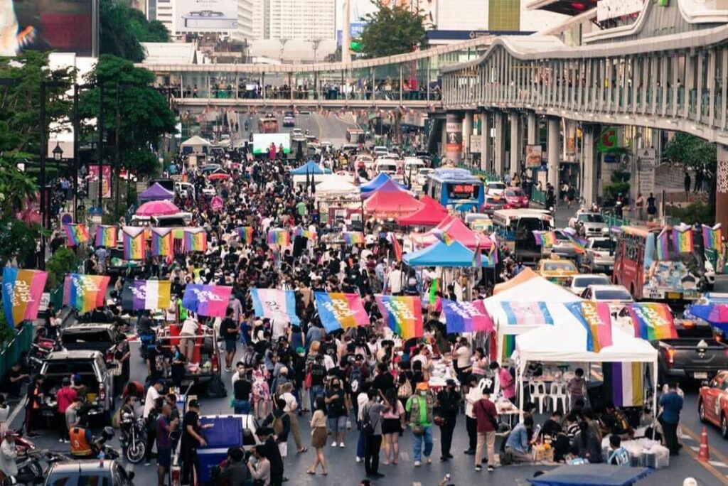 Crowd of diverse individuals celebrating at a colorful Bangkok Pride event, with rainbow flags and vibrant costumes showcasing LGBTQIA+ community spirit.