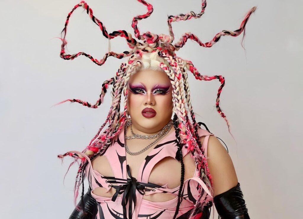Too Calderone confidently posing for the camera in a bold pink and black ensemble, with a unique and imaginative hairstyle that grabs attention.