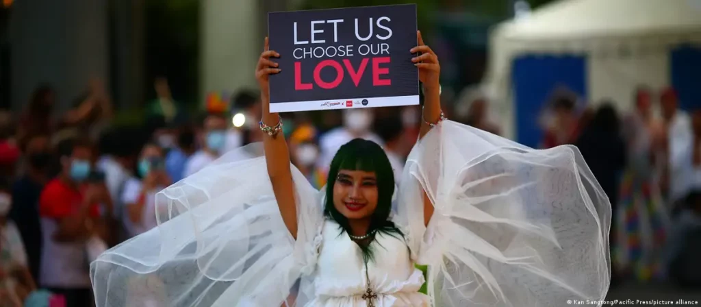 Image of a woman in a wedding dress costume with vibrant green hair, confidently walking on a rainbow flag. She is holding a poster that reads 'let us choose our love', prominently featuring HER logo as a symbol of alliance. The scene conveys a powerful message of love and inclusivity.