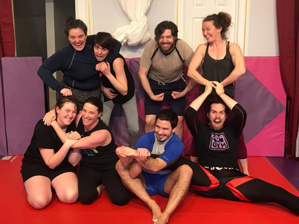 A diverse group of LGBTQIA+ individuals proudly smiling and flexing their muscles inside Pop Gym, a U.S.-based inclusive fitness center promoting a supportive and welcoming environment for all.