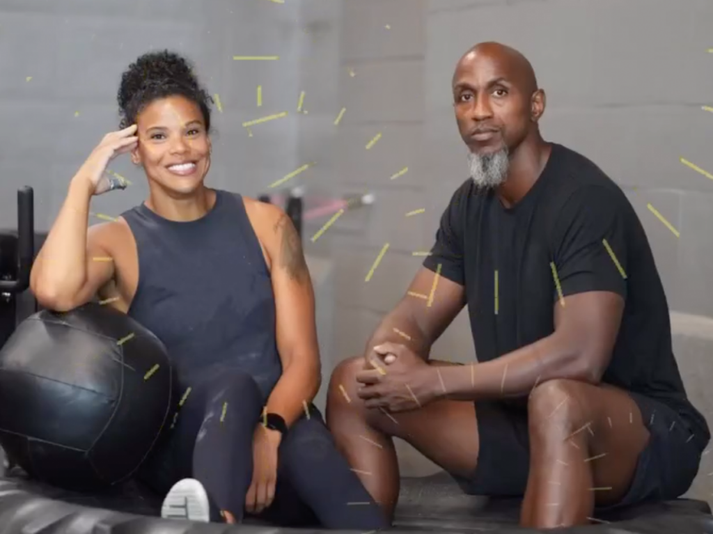 Elisabeth Akinwale, a former professional CrossFit athlete now a social worker, and Kevin Brathwaite, an experienced strength coach, are seated on large exercise balls at 13th Flow, an LGBTQIA+ friendly and inclusive fitness gym in the U.S. They are smiling and looking directly at the camera, symbolizing the welcoming and diverse atmosphere of the gym.