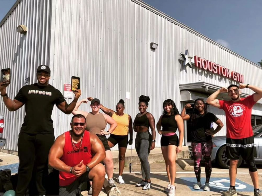 A group of LGBTQIA+ individuals, radiating confidence and inclusivity, stand proudly in front of Houston Gym. They are smiling and flexing their muscles, showcasing their fitness achievements. This image represents one of the top LGBTQIA+ gyms in the U.S., promoting an inclusive fitness environment.
