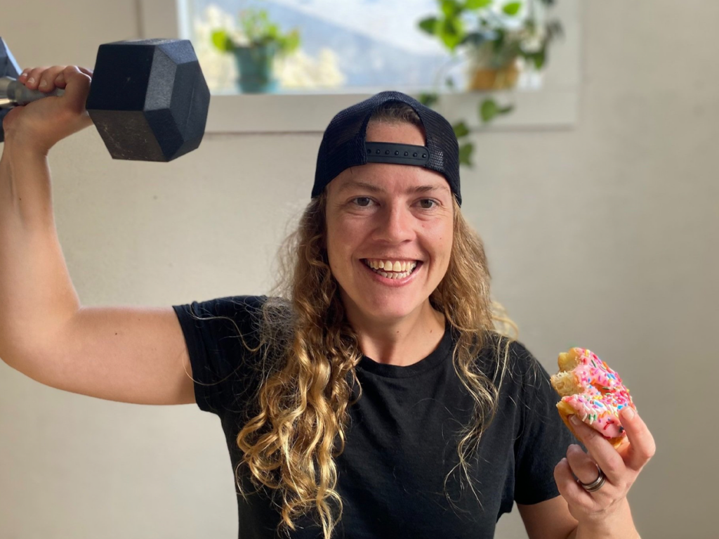 An LGBTQIA+ woman at No Judgement Fitness in the U.S., embodying the spirit of Inclusive Fitness. She is joyfully balancing her fitness and self-care, holding a weight in one hand and a donut in the other, with a bright smile on her face, symbolizing a harmonious blend of health and happiness.