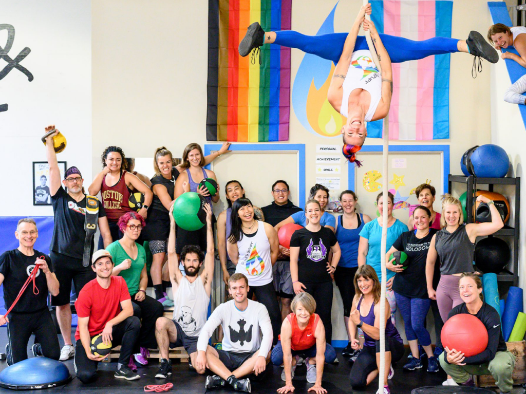 An inclusive and diverse group of LGBTQIA+ individuals posing for a group photo at Strive & Uplift, a gym promoting Inclusive Fitness in the U.S. They are smiling at the camera with a backdrop of rainbow flags. One person is playfully hanging from a piece of gym equipment on the ceiling, adding a fun and dynamic element to the scene.