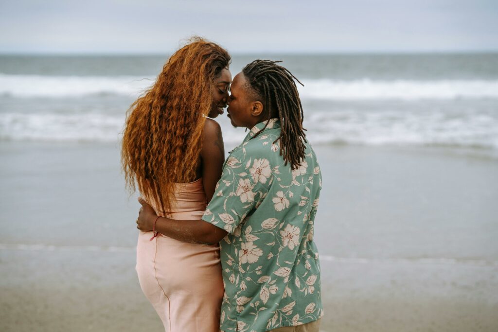 Two lesbians, standing near the shoreline on a beach, share an intimate moment, about to kiss
