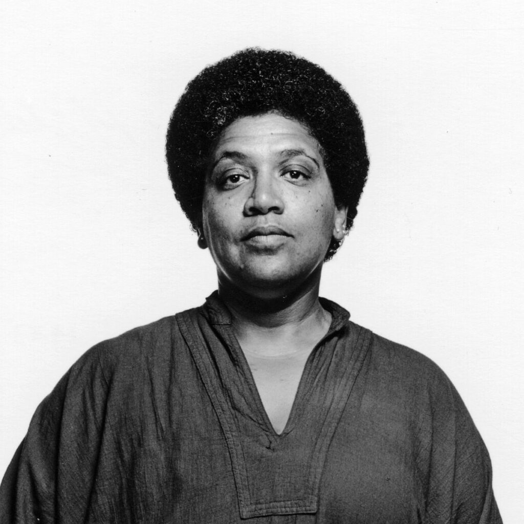 Black and white portrait of Audre Lorde, a Black poet, activist, and feminist, looking confidently at the viewer.