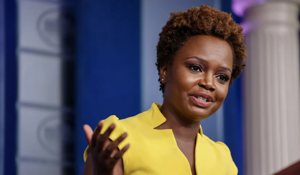 Karine Jean-Pierre, a Black woman with short hair, stands at a podium wearing a yellow dress. She gestures with her hands while speaking confidently to an audience out of frame.