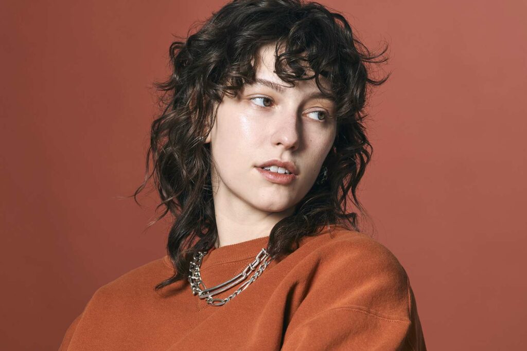 Musician King Princess confidently poses against a vibrant orange background, wearing a casual orange shirt.