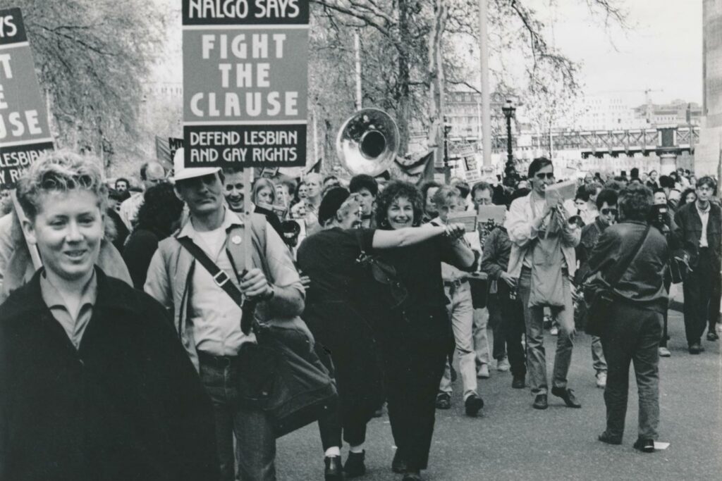 A historic image from 1988 showing demonstrators carrying placards, accompanied by a brass band, in a Stop the Clause protest march. One sign reads 'NALGO says fight the clause. Defend Lesbian and Gay Rights.' This demonstration, opposing the introduction of Section 28, occurred on April 30th, 1988, at Victoria Embankment in London.