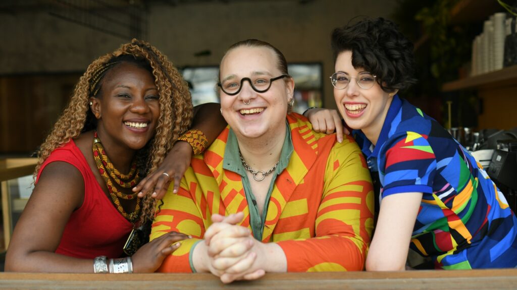 Three vibrant individuals seated, overseeing The Common Press, previously known as Glass House, a sanctuary for London's LGBTQ+ and BIPOC communities.