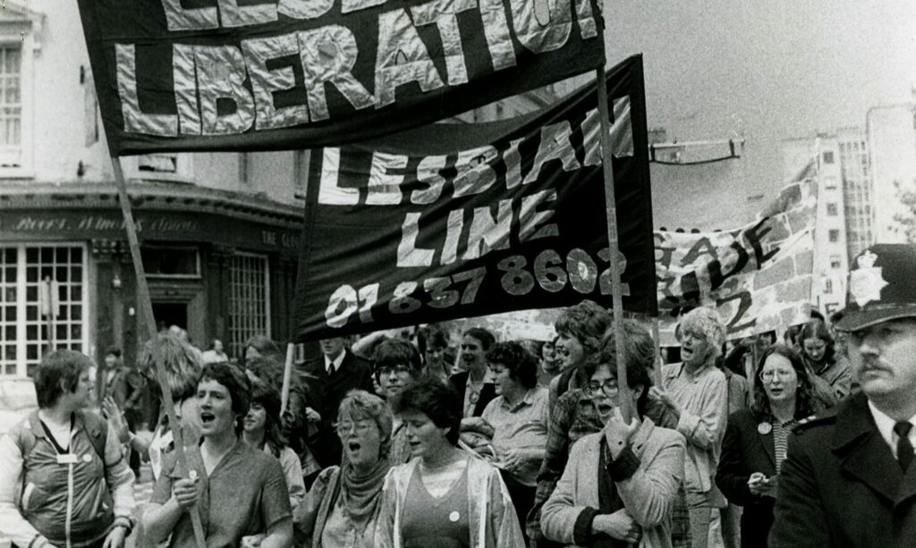 Group of queer individuals and lesbians marching at the Lesbian March in London, 1982. Two people hold a sign that reads 'Lesbian Line 018378602' while a police officer stands nearby.