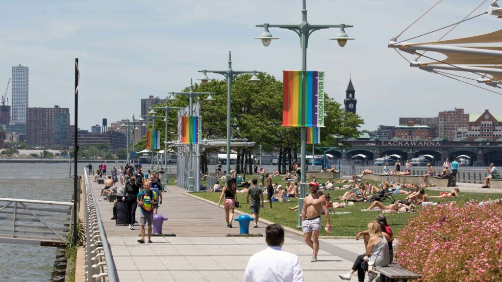 Vibrant scene at Christopher Street Pier: Queer individuals stroll and picnic under the daytime sun, while Pride flags adorn light poles.