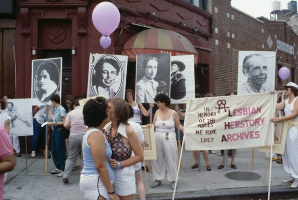 Two women joyfully embrace and kiss, holding up a banner for the Lesbian Herstory Archives in front of the Duchess, a popular lesbian bar in Greenwich Village during Pride in 1982.