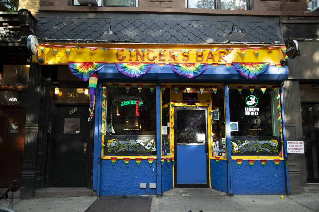 Exterior of Ginger's Bar - Park Slope in New York, adorned with vibrant pride flags and dark blue and yellow walls.