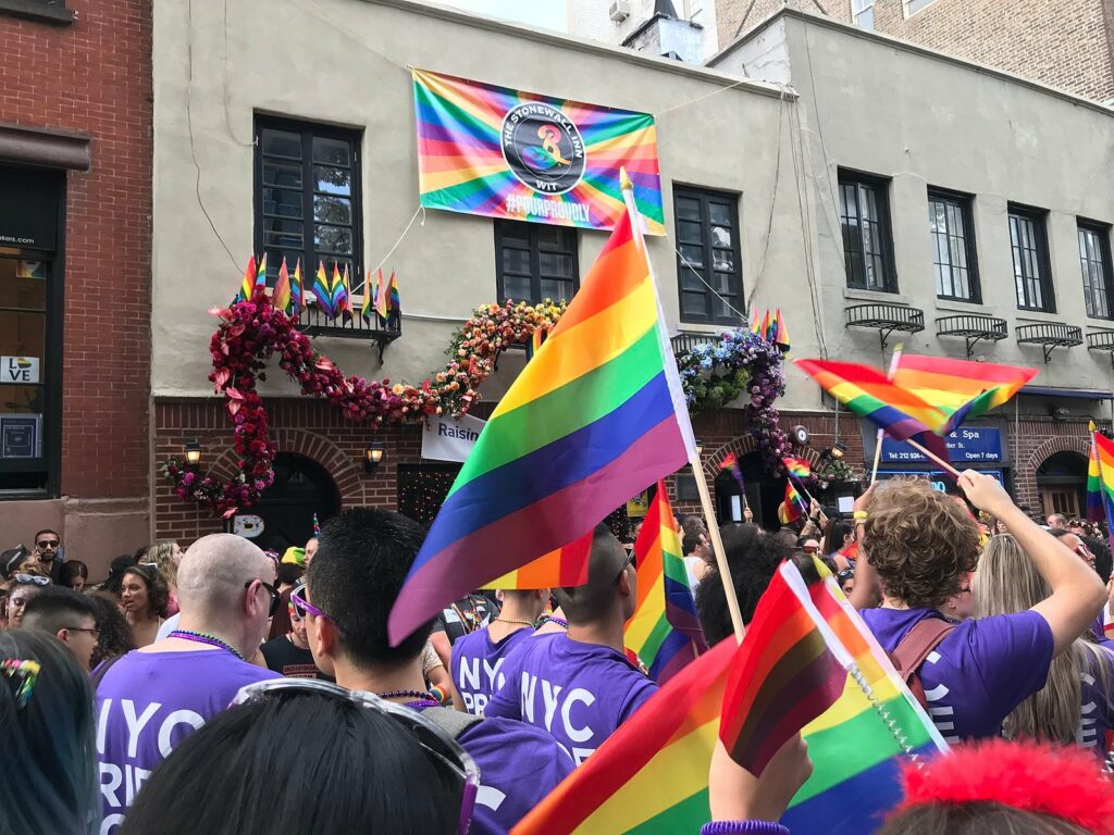 A vibrant Pride Parade outside the historic Stonewall Inn in Greenwich Village. People are dressed in purple NYC Pride shirts and energetically waving rainbow pride flags