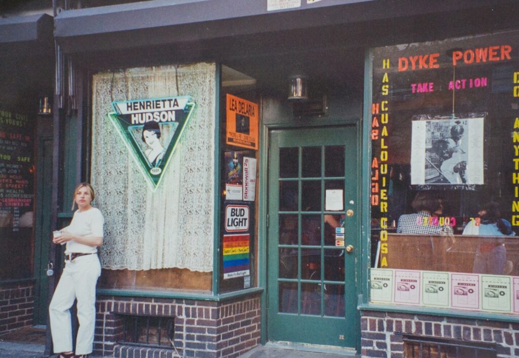Outside of Henrietta Hudson, the nation's oldest lesbian bar, featuring a queer woman in a white t-shirt and light jeans standing in front. The bar's window displays text reading 'DYKE POWER TAKE ACTION' with light-colored flag paper stuck to the window.