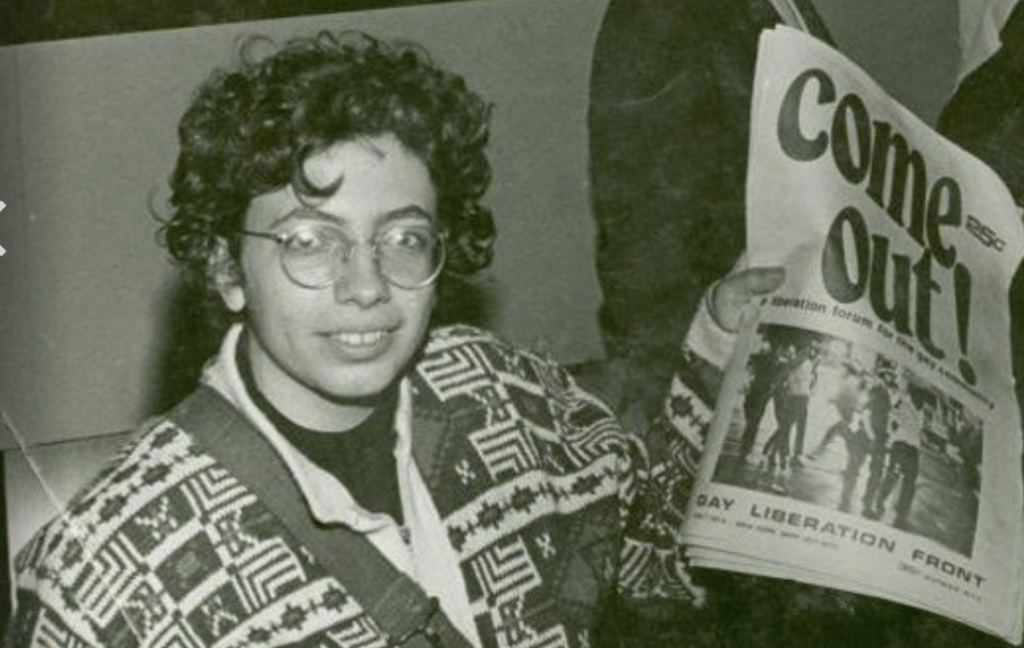 Martha Shelley, a lesbian activist, holds a newspaper with the headline "Come out!"