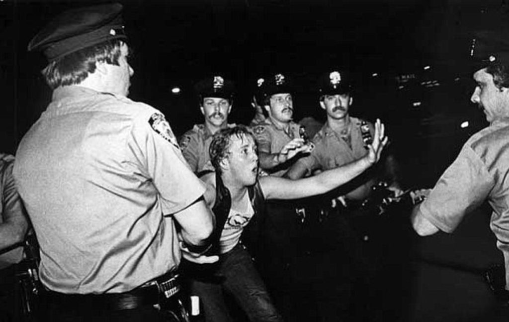 A historic photo depicting a police raid on a bar, dated June 28th, 1969. In the foreground, a queer woman is seen defending herself amidst the chaos.