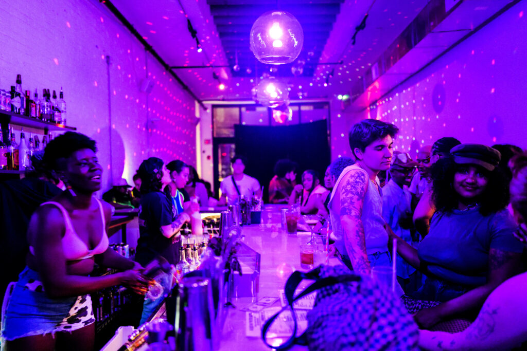 Scene at The Bush bar: A vibrant atmosphere with purple lighting illuminating a diverse crowd, predominantly queer women and lesbians engaged in lively conversations. Smiling bartenders make drinks behind the counter.