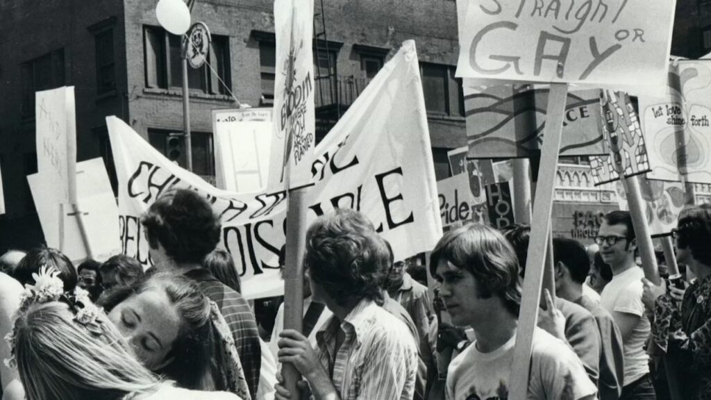Two women, both queer, share a kiss. In the background, a crowd marches, celebrating the first Pride march in NYC on June 28, 1970.