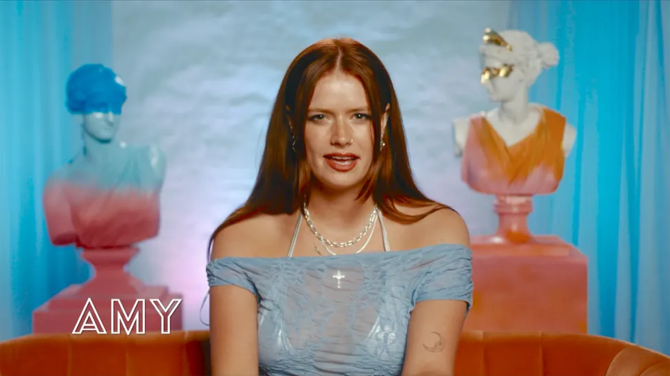 Portrait of Amy, a 24-year-old woman (She/Her) from Surrey, living in London. She wears a see-through blue shirt with a white bar. A cast member from BBC's 'I Kissed a Girl' TV show.