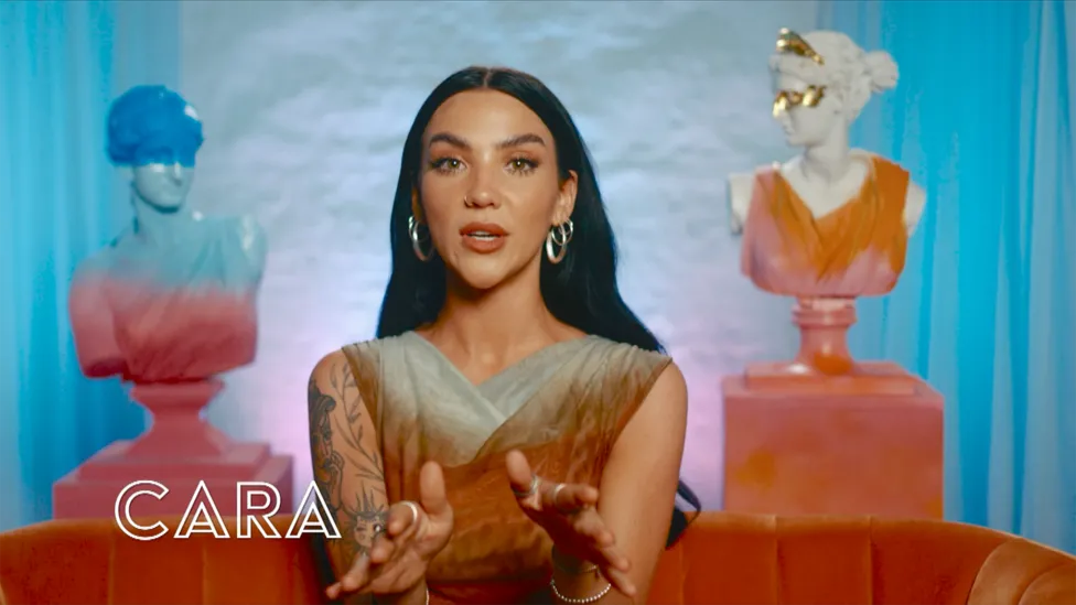 Cara, 25 (She/Her), a cast member from BBC's I Kissed a Girl TV show, wearing a beautiful grayish dress. She proudly displays tattoos on her right arm and wears earrings. Originating from Ballycastle, Northern Ireland, Cara works as a Support Worker, Aesthetician, and Lash Tech.