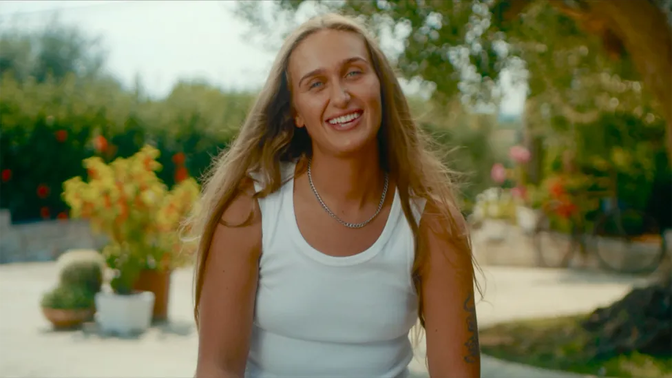 Eva 22 (She/Her), a cast member from BBC's I Kissed a Girl TV show, wearing a white top and smiling. She stands in front of beautiful trees and flowers. Eva 22 is portrayed as a fashionable graduate from Belfast.