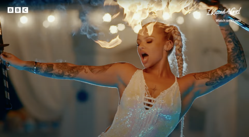 Meg, 24 (She/Her), from Goole, West Yorkshire, a fire breather on BBC's I Kissed a Girl TV show. She stands confidently in a white dress, performing fire breathing while dancing provocatively among other cast members, exuding sexiness.