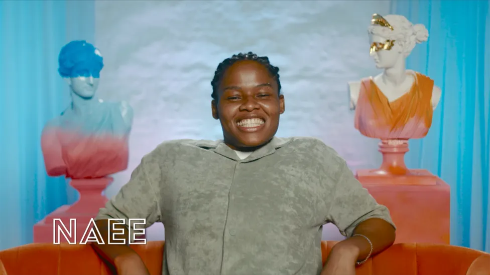 Naee, a 25-year-old woman (She/Her), sits confidently, wearing a light green shirt and smiling. She is a cast member in BBC's 'I Kissed a Girl' TV show. Naee is an engineer from London.