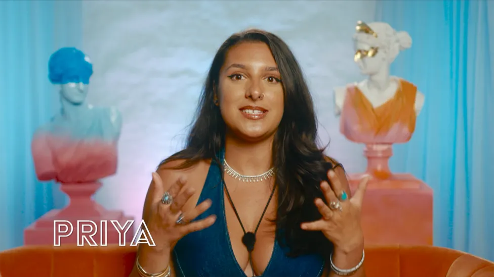 Priya, a 23-year-old woman with dark hair, is gesturing with both hands as she explains something. She wears a stylish dark blue dress, accessorized with rings and a white necklace. She is from Newport, South Wales, and works in hotel customer service. (She/Her)