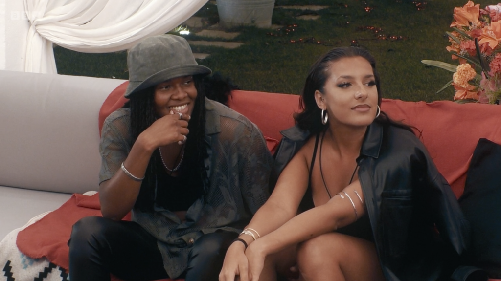 Priya and Naee from I Kissed A Girl tv show, a Match Made in Masseria, are sitting on a sofa enjoying a conversation. They are about to be told that they've been talking behind Hannah's back.