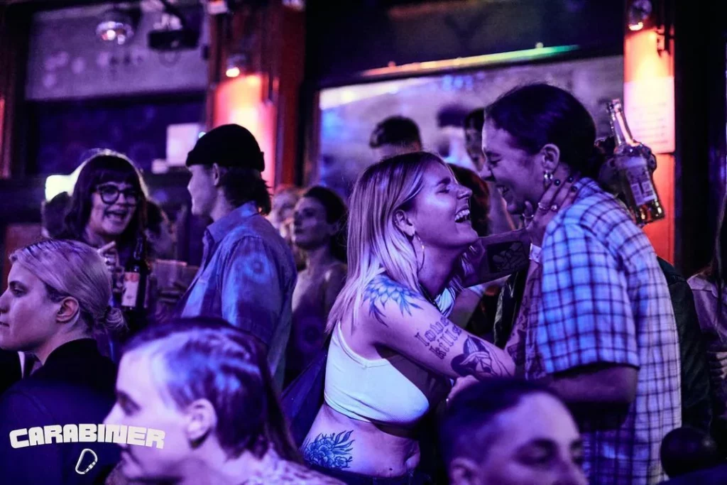 A lesbian couple about to kiss while other sapphic individuals chat at The Big Queer After Party hosted by Big Queer Picnic, part of London Pride events 2024.