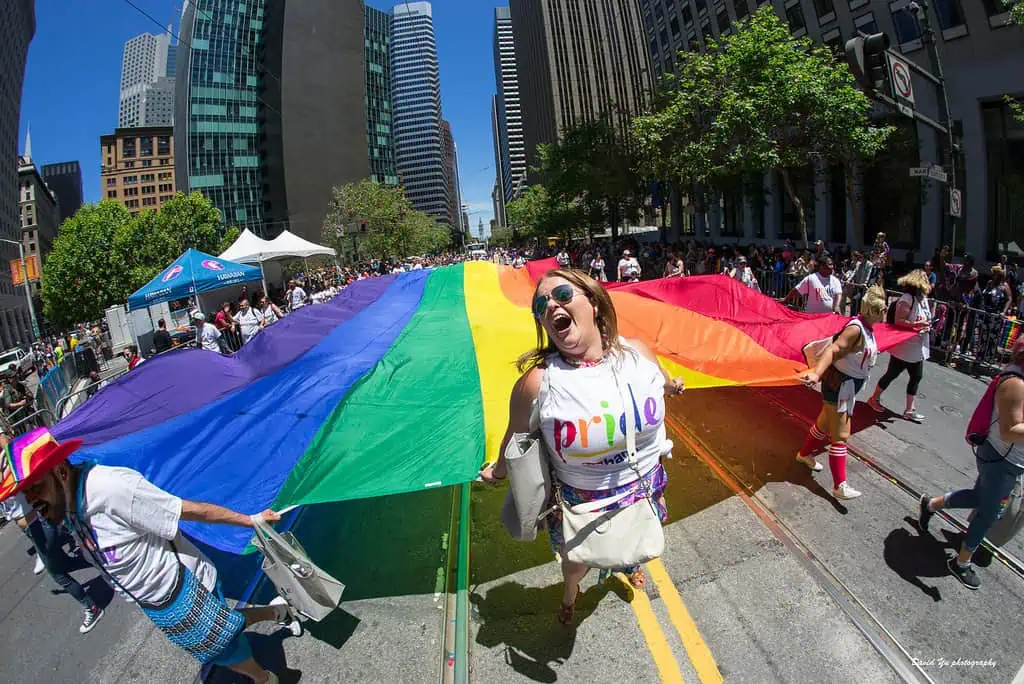 Group of diverse LGBTQ individuals proudly holding a large rainbow flag at the SF Pride Parade 2023, representing unity and celebration.
