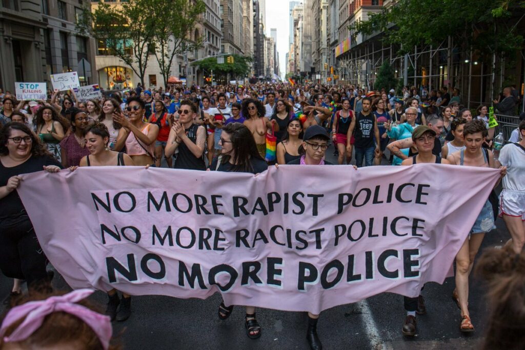 A diverse crowd marches through the streets of New York City during the Dyke March, carrying a large banner that reads 'NO MORE RAPIST POLICE. NO MORE RAPIST POLICE. NO MORE POLICE.' This scene captures the spirit of activism and solidarity at the New York Pride.