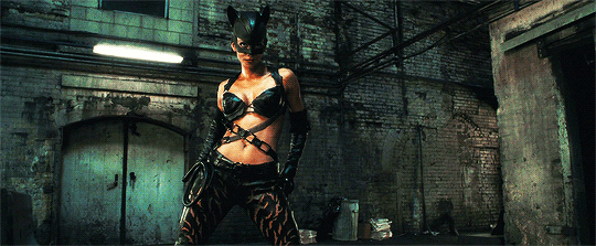 Halle Berry as Catwoman, dressed in a revealing, black leather outfit with a cat mask, playfully handling a gun in a confident pose.