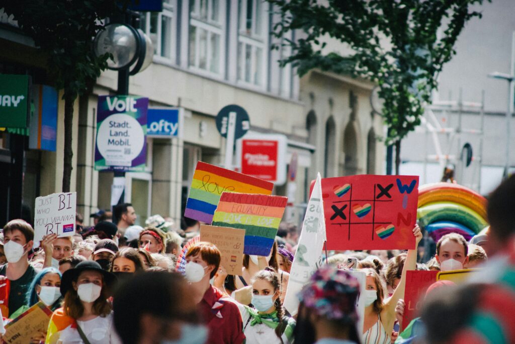 A group of bisexual individuals standing on a street holding a large poster that reads "Just Bi Who Wanna Bi," along with rainbow-themed posters promoting bisexual visibility and equality. The group appears lively and proud, advocating for inclusivity and LGBTQ+ rights.