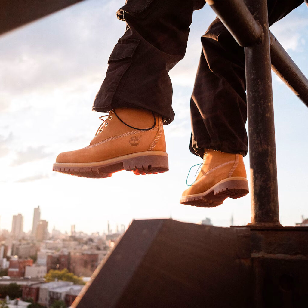 Closeup shot of someone wearing the classic Timberland boots, hanging over a rooftop railing with a city skyline in the background.
