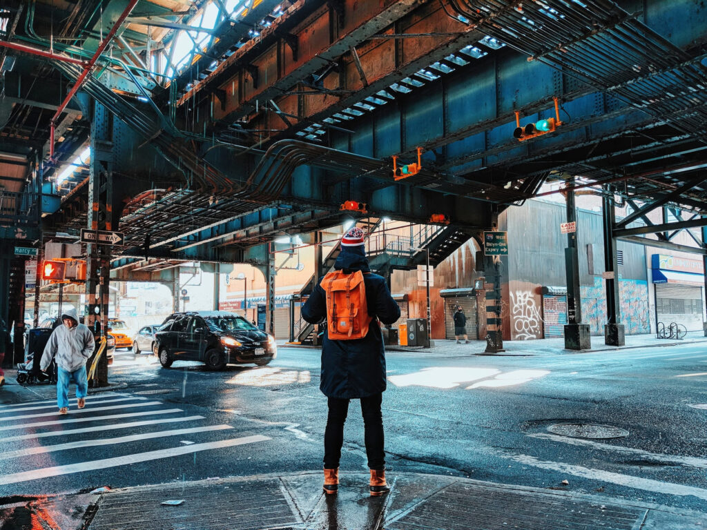 A New Yorker wearing Timberland boots stands on the sidewalk at a Brooklyn intersection underneath above-ground subway tracks.